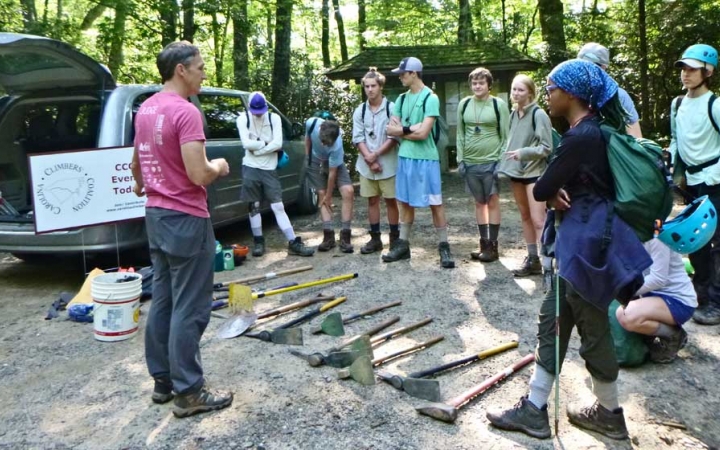 A person speaks to a group of students. There are garden tools laying on the ground between them.